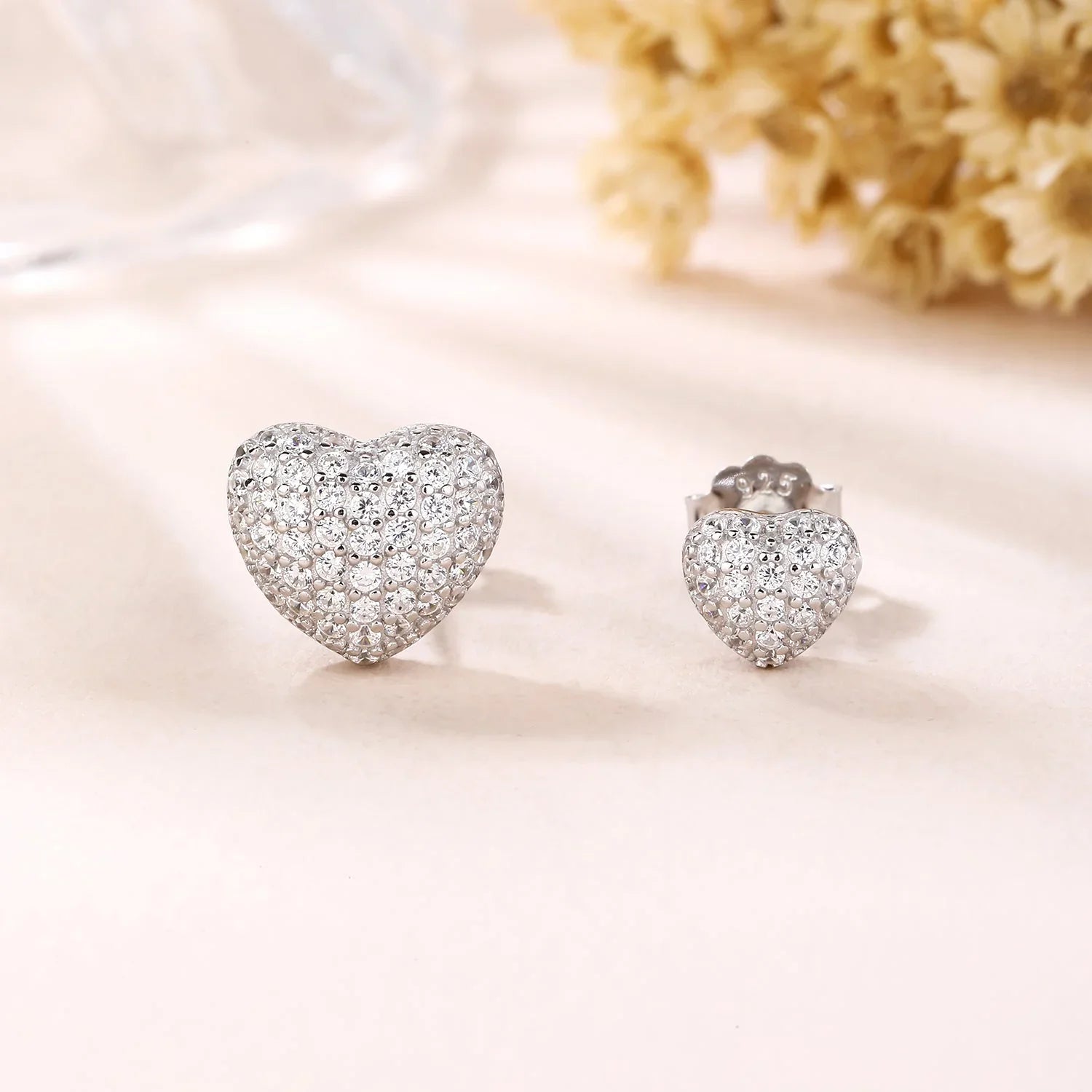 Heart-shaped silver stud earrings with sparkling stones by Moment Jewels on a soft background