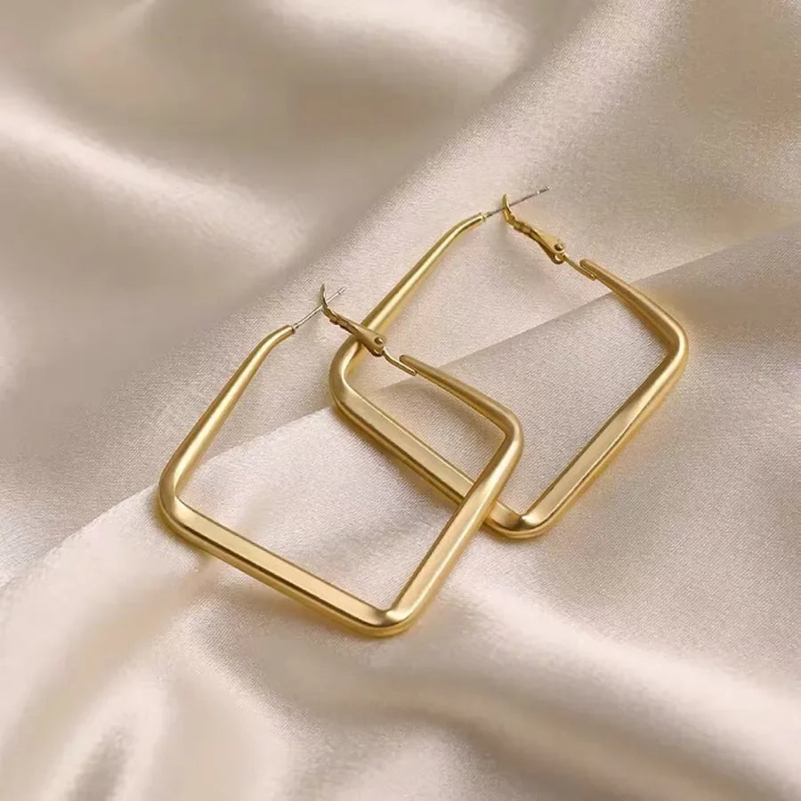 Gold square hoop earrings from Moment Jewels on a soft beige satin fabric background